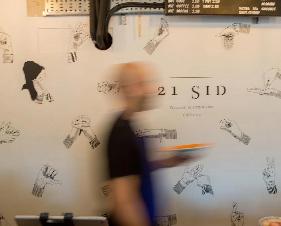 A blur memeber of staff running behind the counter. On their back, we see a white wall with the 21 Sid logo.